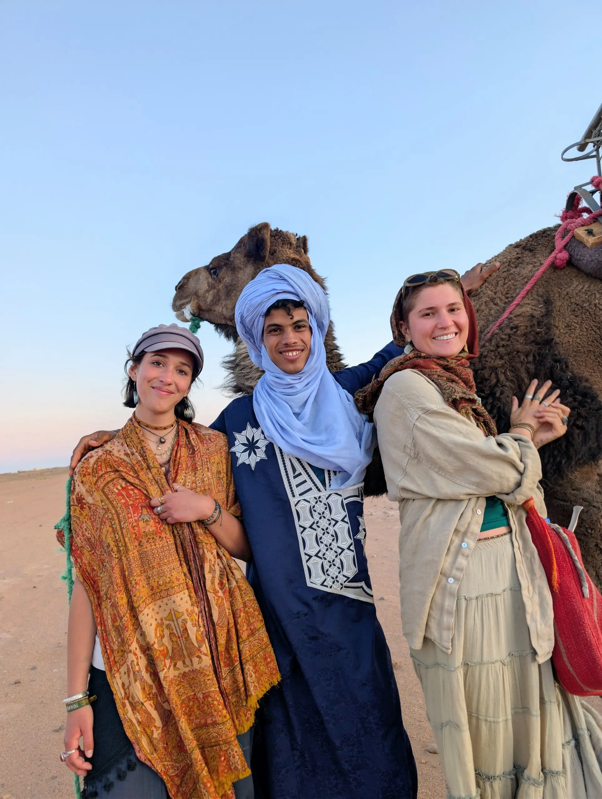 Two travelers posing with a local desert guide and camel during a Sahara tour in Morocco