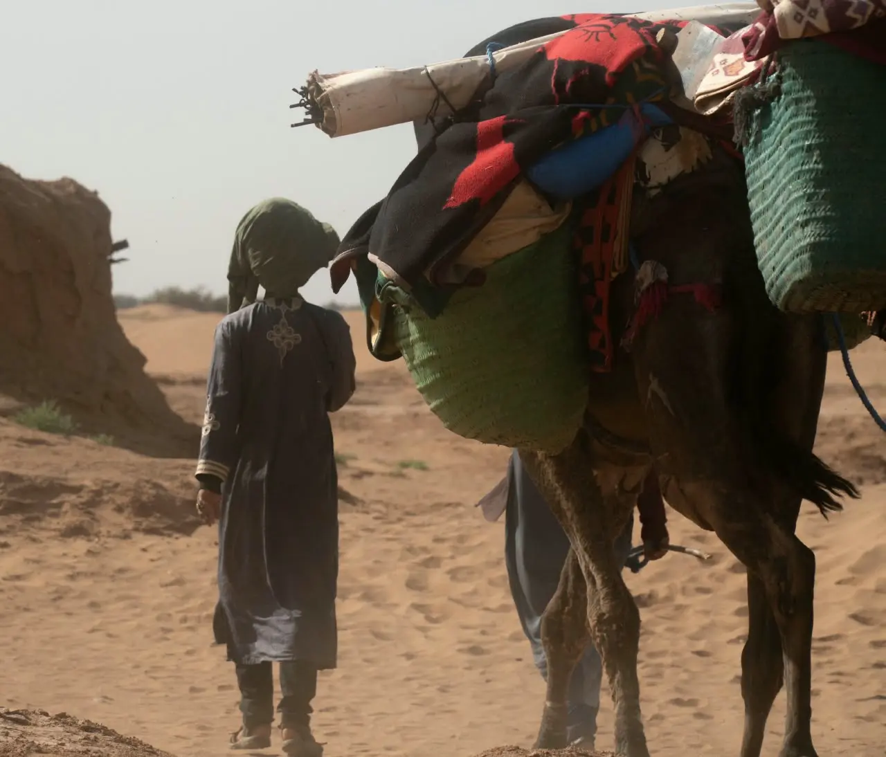 A loaded camel carrying supplies walks through the sandy desert beside a person.