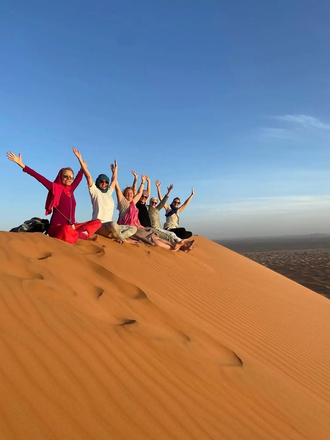 A group of six women travelers sitting in a row on the ridge of a golden desert sand dune, raising their hands in celebration under a clear blue sky