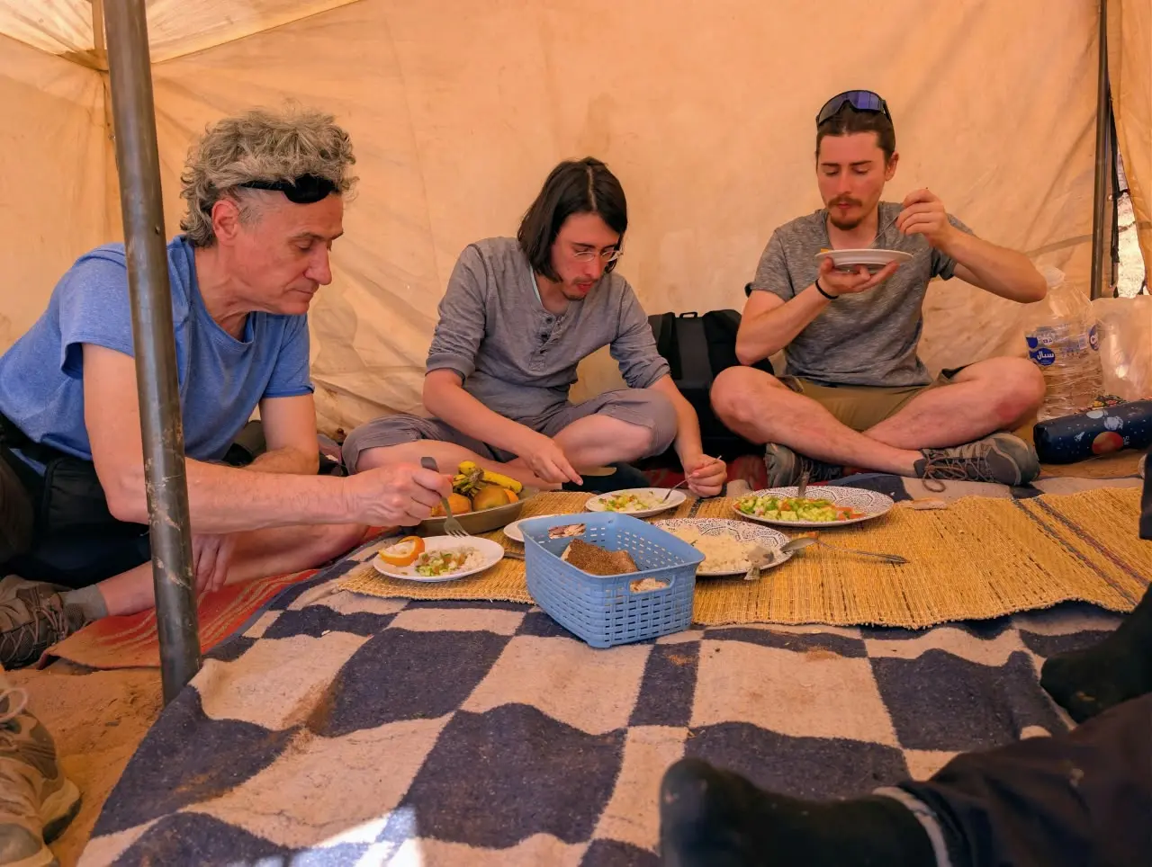 Three travelers share a meal while seated on a patterned rug inside a desert tent.