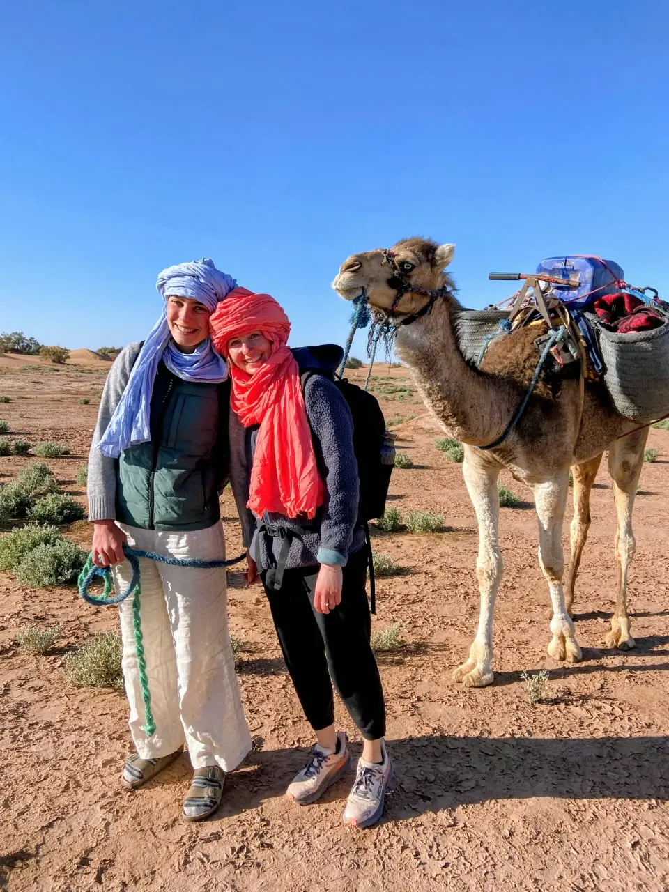 Two travelers stand beside camels on a sandy desert plain beneath a clear blue sky.